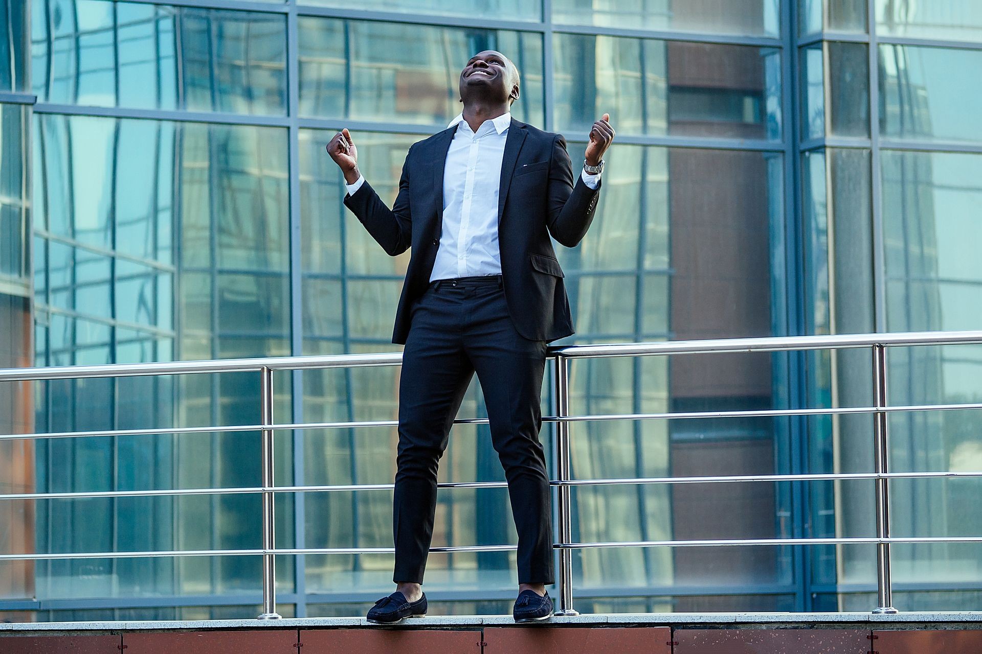 a handsome African American man in a black stylish suit and a white shirt rejoicing in success and thanking God against the background of city landscapes glass business centers. a good deal concept